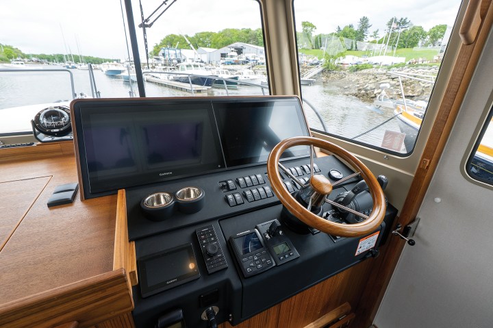 The helm station of a Targa 41 featuring a wooden steering wheel, Garmin dual displays, physical toggle switches, and teak trim.