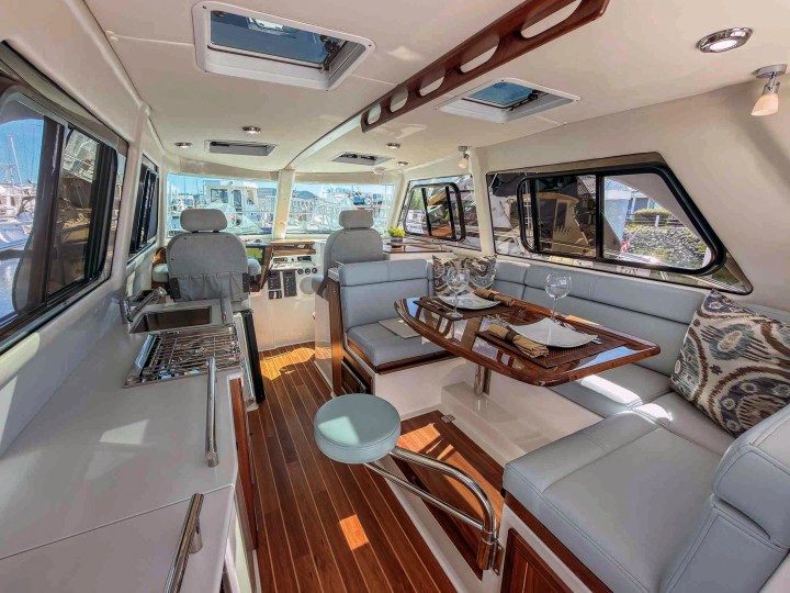 The interior salon of an Aspen C110 featuring a portside linear galley, a wooden dinette table with light grey seating, and dual helm chairs forward.
