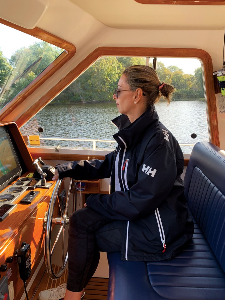 Giselle wearing a navy blue Helly Hansen jacket seated at the wooden indoor helm station of a Grand Banks yacht while steering along a river.