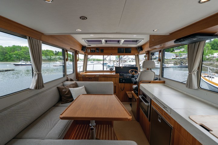 The interior salon of a Targa 41 showing a teak dinette table, grey upholstered seating, large side windows, and a raised galley across from the helm.