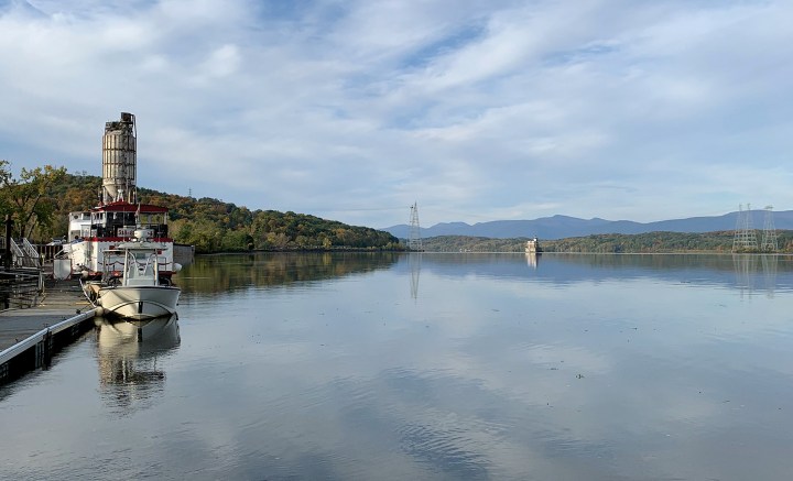 Calm water reflecting cloud-filled skies along the Hudson River with boats docked beside an industrial silo structure and mountains in the distance.