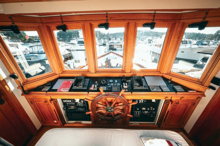 Pilothouse helm of the historic motor yacht Paspatoo featuring a traditional wooden ship's wheel, modern navigation displays, and wide wraparound windows looking out over the marina.