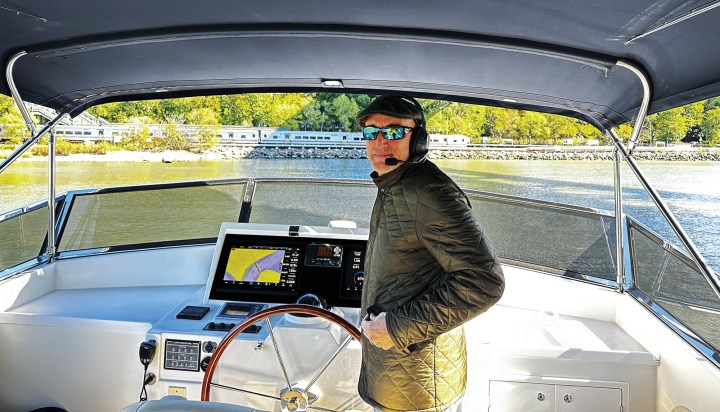 Frank Scavone standing at the flybridge helm of his Grand Banks motor yacht wearing a flat cap and headset, with a passenger train visible along the shoreline behind him.