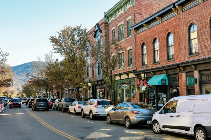 A charming, tree-lined downtown street in Beacon, New York, featuring historic brick storefronts, parked cars, and mountains in the distance.