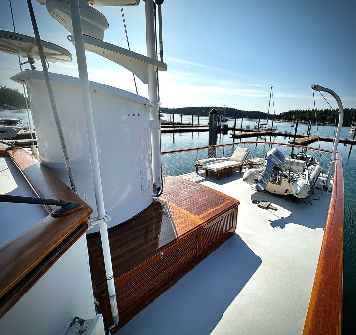 Upper boat deck of the classic motor yacht Paspatoo featuring teak cabinetry, loungers, radar mast, and a stowed tender with crane davit.