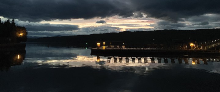 A quiet dock extending into calm waters illuminated by dock lights under a dark, moody sunset sky with dramatic cloud formations.