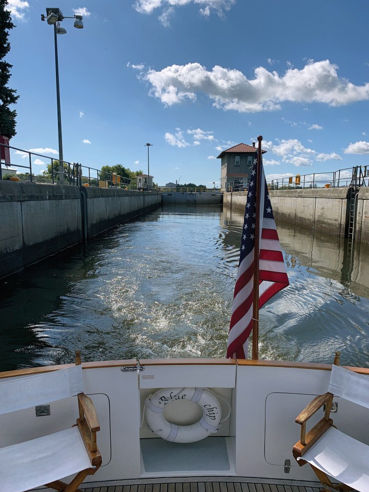 The stern of a yacht named Blue-chip displaying an American flag while entering a concrete canal lock under a bright blue sky.