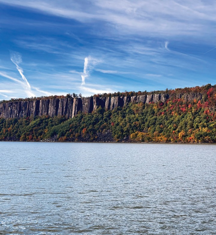 The dramatic basalt cliffs of the Palisades rising above the Hudson River with colorful autumn foliage along the shoreline under a bright blue sky.