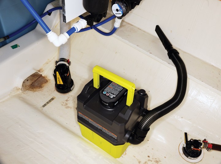 A cordless lime-green and black Ryobi shop vacuum sitting inside a boat bilge compartment alongside plumbing hoses, strainers, and thru-hull fittings.
