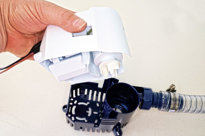 A person holding the white upper float switch and motor housing removed from a blue Rule bilge pump strainer base and discharge hose to inspect for debris.