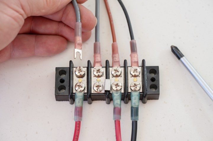 A hand sliding an open flanged fork terminal onto an electrical terminal block wired with marine heat-shrink crimp connectors beside a screwdriver.