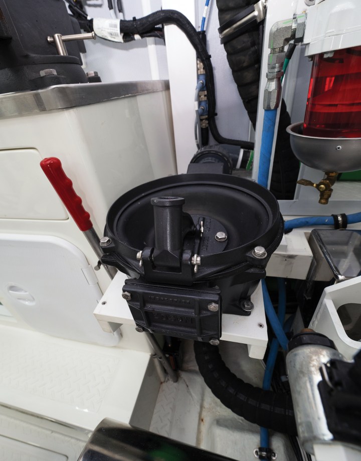 A heavy-duty manual diaphragm bilge pump mounted on a bulkhead inside an engine room featuring a red operating handle and black intake plumbing.