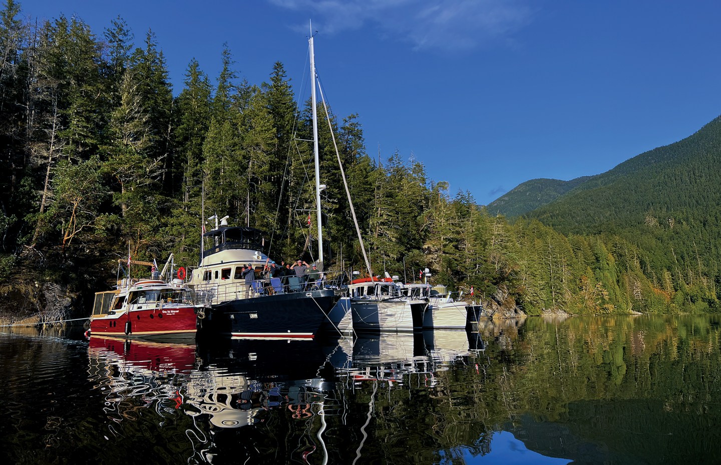 Flotilla of powerboats and a sailboat rafted together in a calm, forested cove during a Pacific Northwest cruising expedition.