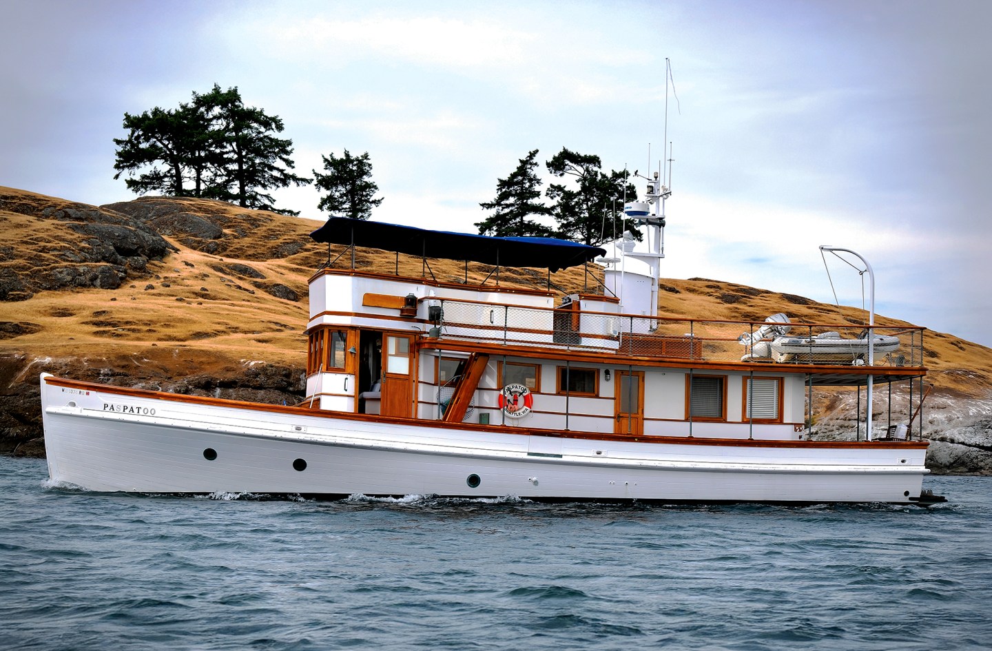 Side profile of the historic 66-foot wooden motor yacht Paspatoo cruising past a grassy shoreline.