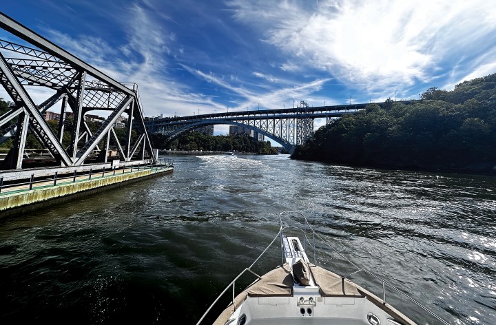View from the bow of a yacht navigating a narrow river passage beneath structural bridges under a blue sky with scattered clouds.