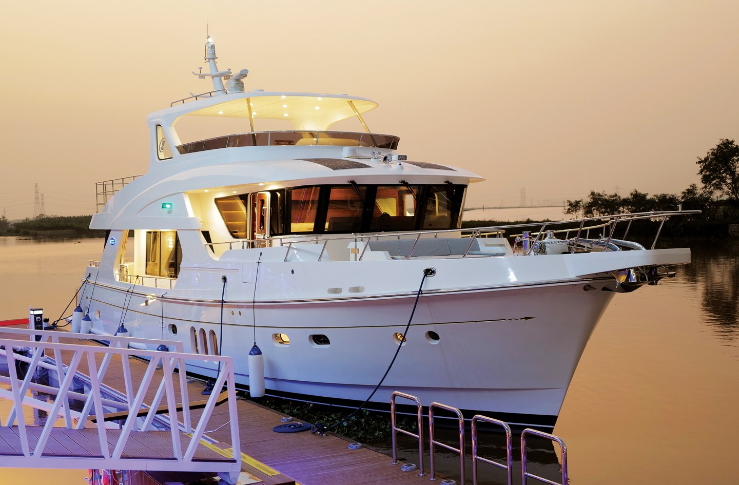 A luxury white Selene motor yacht docked along a pier at dusk with warm exterior lighting illuminated.